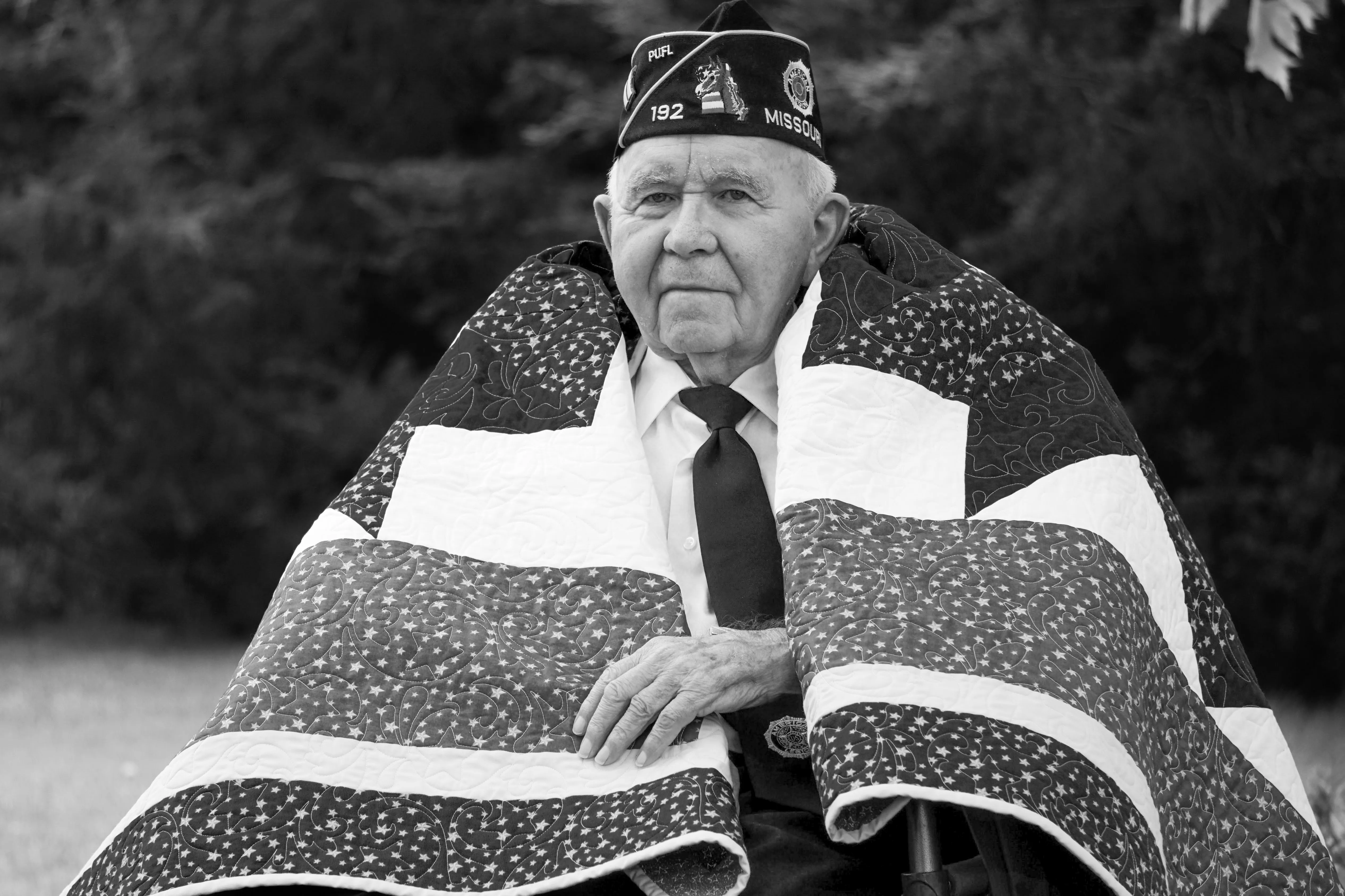 An older man in an American Legion veteran's cap with a red, white, and blue quilt draped over him.